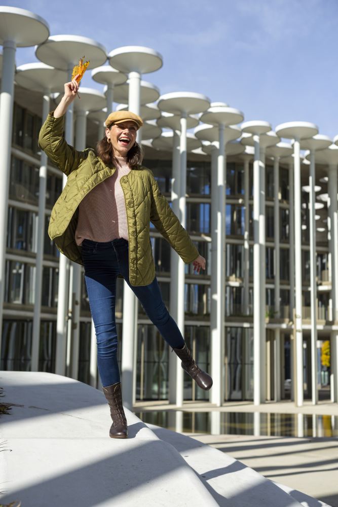 Eine fröhliche Person im ATTRAQT Stretch Nappa Brown von ZAQQ balanciert flexibel auf einem Bein und hält einen Blumenstrauß in der Hand. Sie stehen vor einer Kulisse aus modernen, zylindrischen Architektursäulen an einem hellen Tag.