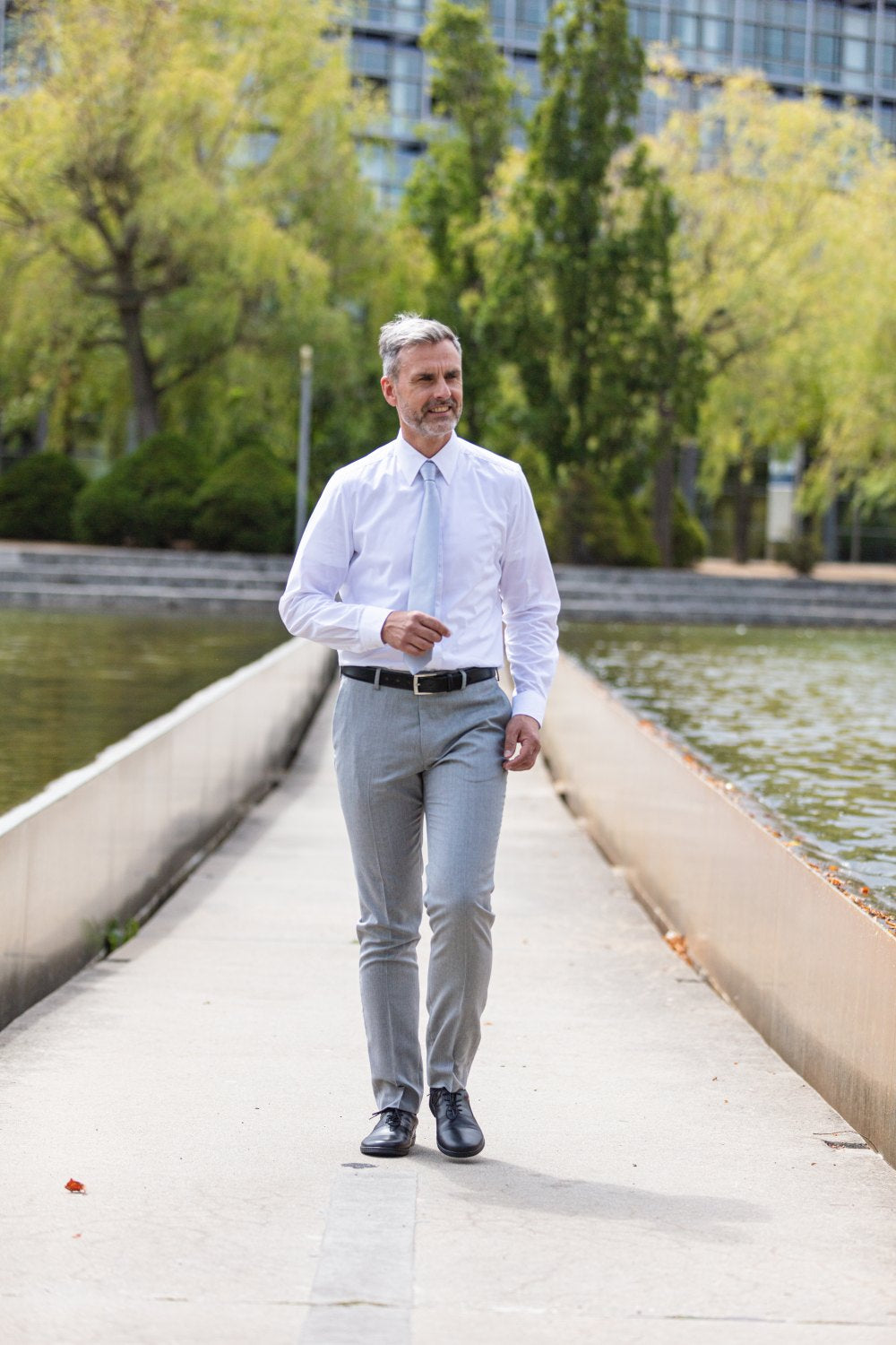 Ein Mann in weißem Hemd und grauer Hose geht selbstbewusst auf einem schmalen Pfad entlang eines Gewässers. Seine Schritte sind durch die eleganten schwarzen APEQ-Schuhe von ZAQQ sicher. Im Hintergrund sind Bäume und ein modernes Gebäude unter einem klaren Himmel zu sehen.