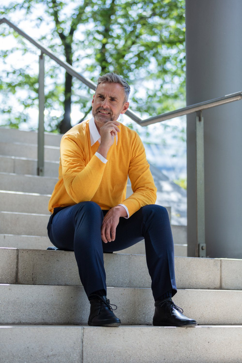 Ein Mann in gelbem Pullover und marineblauer Hose sitzt nachdenklich auf einer Außentreppe und stützt sein Kinn auf seine Hand. Die schwarzen ARQ-Stiefel von ZAQQ vervollständigen seinen Look, umgeben von einem Metallgeländer und üppigen grünen Bäumen im hellen Tageslicht.