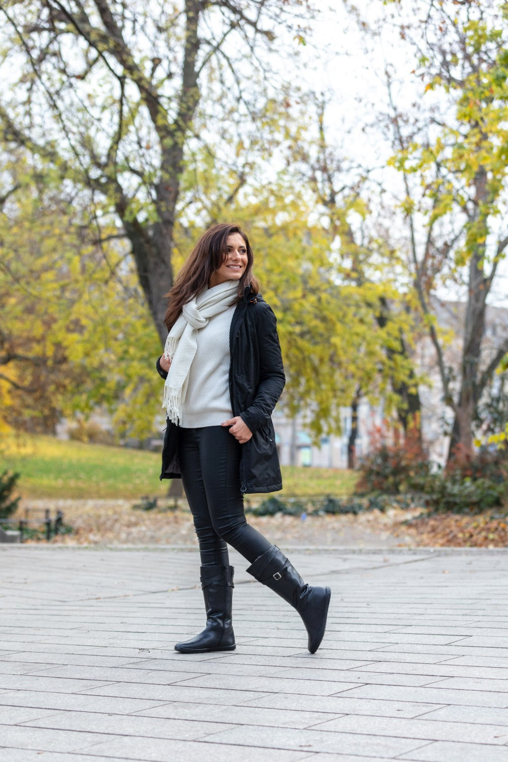 Eine Frau in schwarzem Mantel, weißem Schal und schicken ZAQQ ATTRAQT Winter Black Boots aus Ziegennappaleder schlendert auf einem gepflasterten Weg durch einen Park. Im Hintergrund sind herbstliche Bäume mit gelben Blättern zu sehen, die die lässige, herbstliche Atmosphäre verstärken.