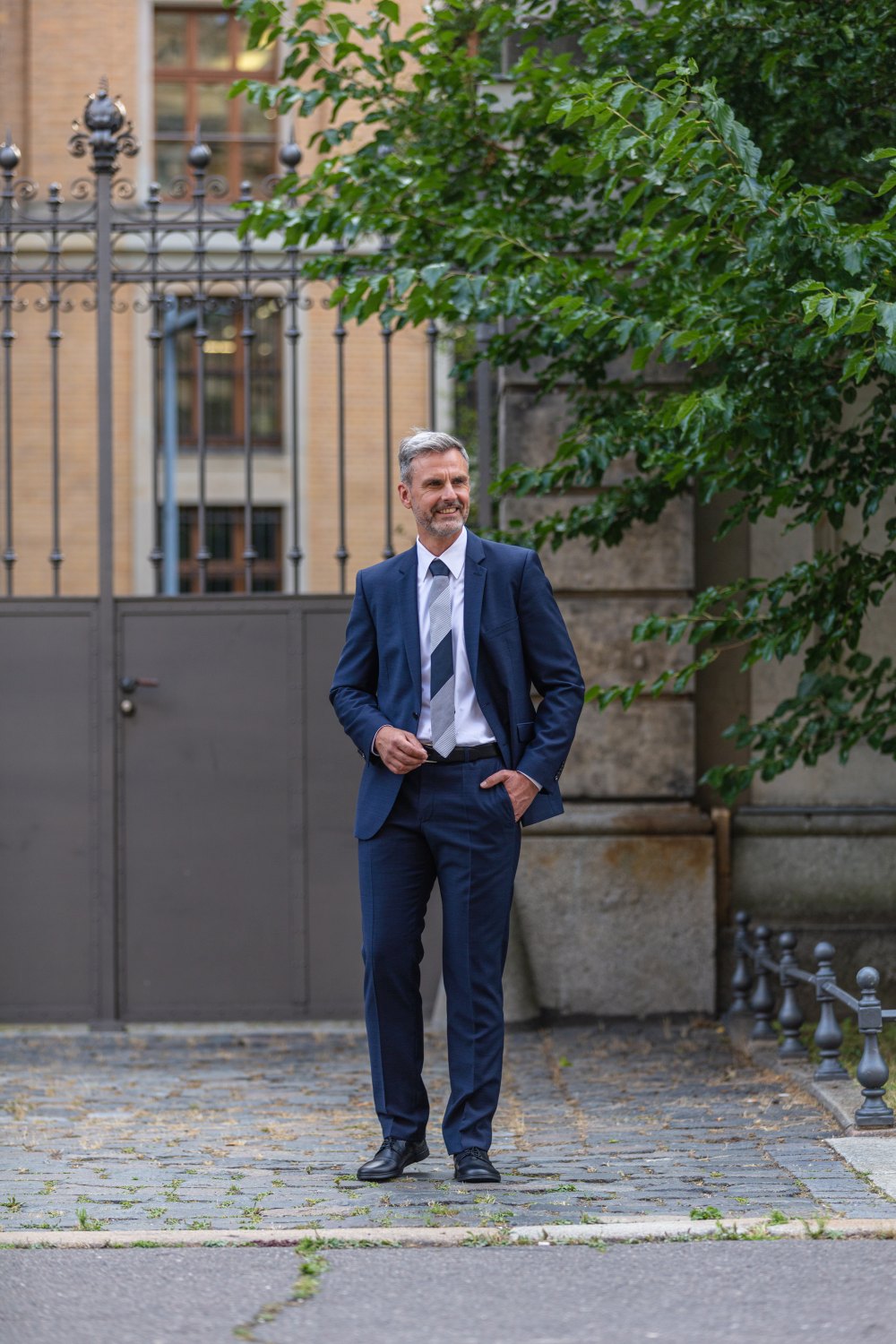 Ein Mann in einem marineblauen Anzug steht selbstbewusst auf einem gepflasterten Weg im Freien, die Hand in der Tasche. Er trägt eine Krawatte und schicke schwarze Oxfords BRIQ Captoe von ZAQQ. Hinter ihm sind ein Metalltor und Grünflächen zu sehen, im Hintergrund ist ein Gebäude zu sehen.