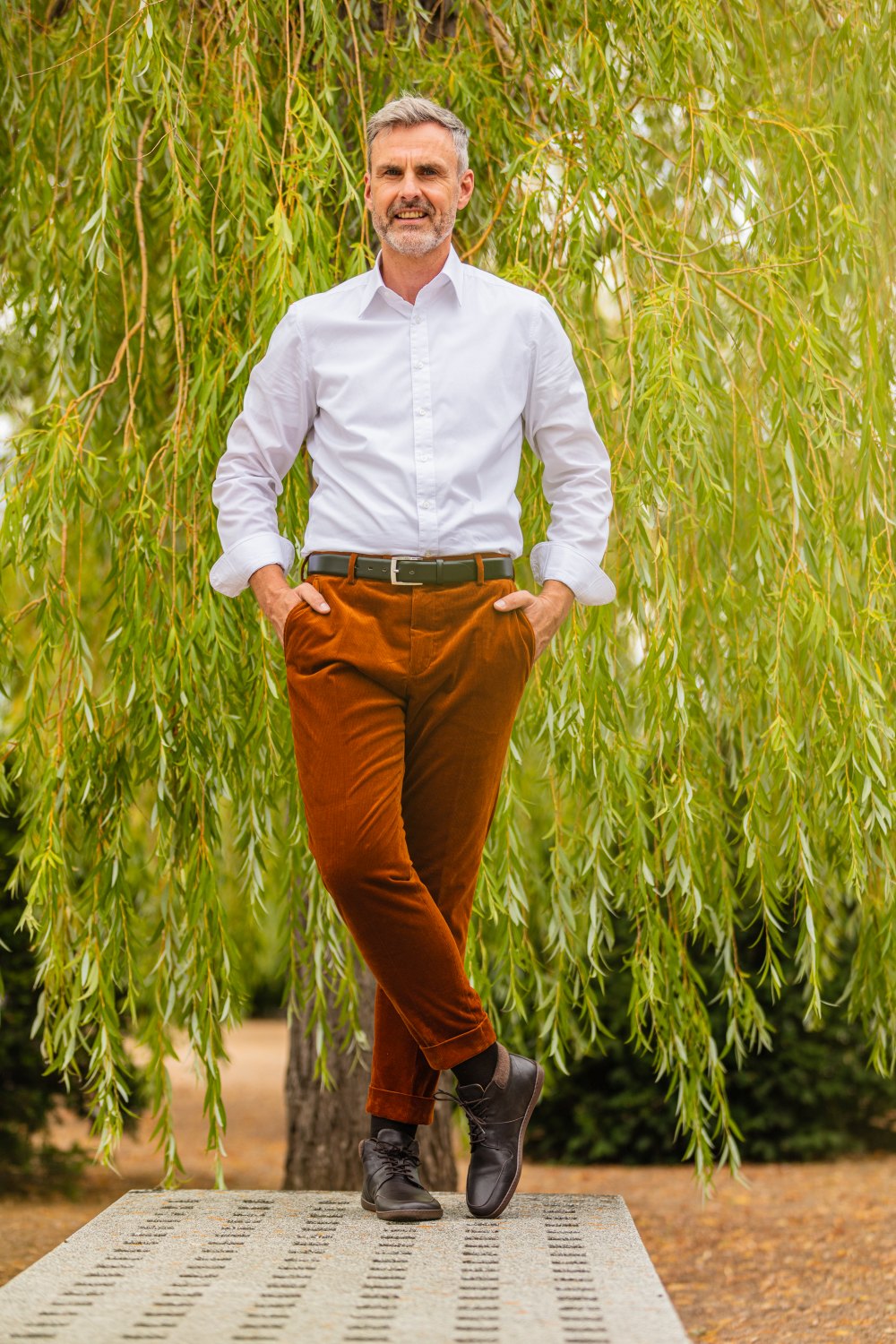 Ein Mann mit grauem Haar und Bart steht selbstbewusst auf einer Steinbank. Er trägt ein weißes Hemd, braune Hosen und dunkle NEWQUAY Brown-Stiefel von ZAQQ. Ein üppiger grüner Trauerbaum dient als Hintergrund.