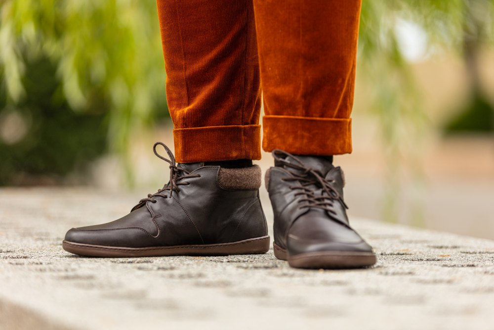 Eine Person, die dunkelbraune NEWQUAY High-Top-Schuhe von ZAQQ und hochgekrempelte Hosen in knalligem Orange trägt, steht auf einer Betonfläche. Die Schuhe haben dunkle Schnürsenkel und einen gepolsterten Knöchelkragen. Der verschwommene Hintergrund aus grünem Laub verleiht dem stilvollen Ensemble einen Hauch von Natur.