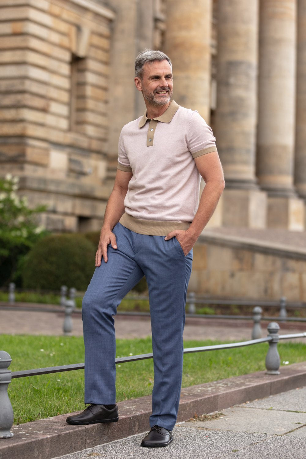 Ein Mann mit grauem Haar und Bart steht selbstbewusst im Freien. Er trägt ein hellrosa Poloshirt und eine blaue Hose, die den Business-Casual-Look perfekt einfangen. Er lächelt leicht, die Hand auf der Hüfte, während ein historisches Gebäude mit großen Säulen den Hintergrund bildet. Seine PEAQ Dark Brown-Schuhe von ZAQQ verleihen ihm einen zusätzlichen Hauch von Stil.