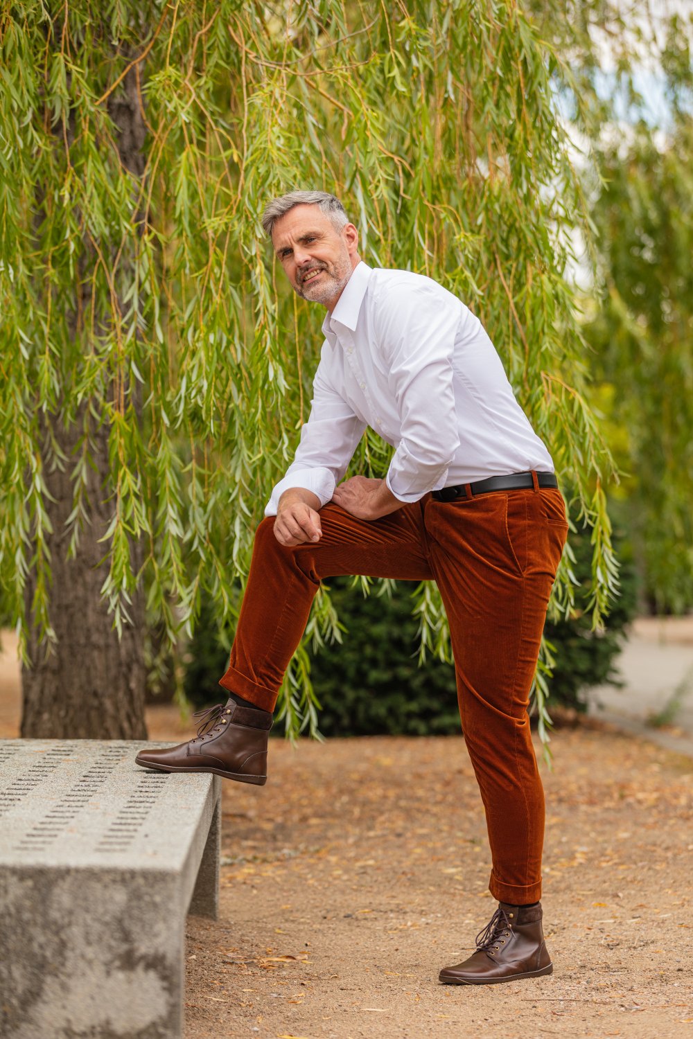 Ein Mann mit grauem Haar, der ein weißes Hemd und braune Hosen trägt, posiert mit einem Fuß auf einer Bank im Park und präsentiert seinen ZAQQ QORBY Brown-Stil. Hohes Grün und Bäume bilden den Hintergrund, während der Boden mit brauner Erde bedeckt ist.