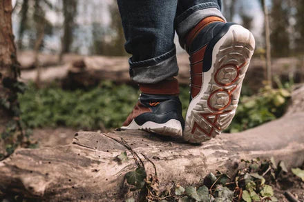 Nahaufnahme einer Person in Wanderschuhen, die auf einem umgestürzten Baumstamm steht. Die Sohle eines Schuhs ist sichtbar und zeigt das Markenlogo. Die Person trägt dunkle Jeans und ist von einer Waldkulisse mit viel Grün und verschwommenen Bäumen im Hintergrund umgeben.