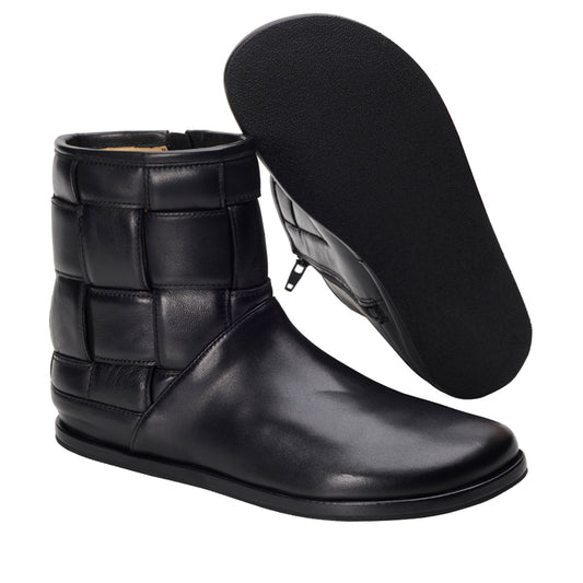 Wir präsentieren die LUQAS Wide Black von ZAQQ, ein elegantes Paar schwarzer Stiefel aus Nappaleder mit gepolsterten quadratischen Nähten und einer flachen, barfuß-inspirierten Sohle. Ein Stiefel steht aufrecht, während der andere geneigt ist und einen Blick auf die sorgfältig gefertigte Sohle bietet.