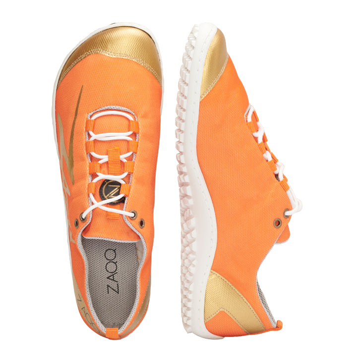 Wir präsentieren den SQIM Orange Gold, ein Paar Laufschuhe von ZAQQ. Diese Schuhe haben goldene Akzente und sind aus Recyclingmaterialien gefertigt. Sie haben weiße Laufsohlen und Schnürsenkel. Der Markenname ZAQQ ist elegant auf der Innensohle eines Schuhs abgebildet. Sie sind für höchsten Tragekomfort konzipiert und werden nebeneinander abgebildet, wobei einer aufrecht steht und der andere auf der Seite liegt.