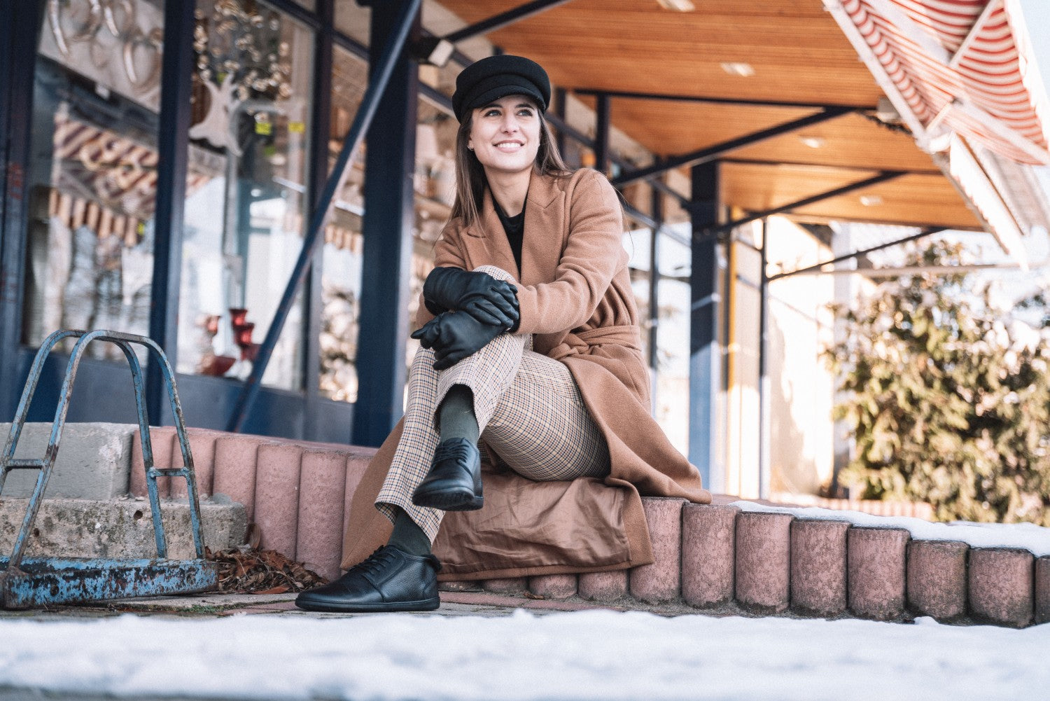 Eine Frau sitzt draußen auf einem Ziegelsteinsims. Sie trägt einen braunen Mantel, karierte Hosen, schwarze Handschuhe und eine schwarze Mütze. Sie lächelt und stützt ihre Arme auf ihre Knie. Die Szenerie umfasst ein Gebäude mit großen Fenstern und etwas Schnee auf dem Boden. Der Himmel ist klar und sonnig.