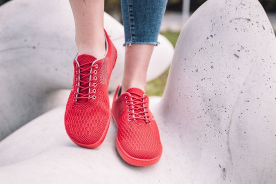 Nahaufnahme einer Person, die leuchtend rote Turnschuhe mit Schnürsenkeln auf einer glatten, abstrakten weißen Skulptur trägt. Die Person trägt Blue Jeans und im Hintergrund ist ein verschwommener Hauch von Grün zu sehen, der eine Umgebung im Freien suggeriert.