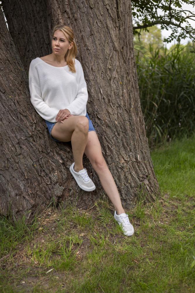 Eine Person mit langen Haaren sitzt an einem großen Baumstamm gelehnt. Sie trägt einen weißen Pullover, Jeansshorts und elegante weiße ALIQANTE-Sneaker von ZAQQ aus geschmeidigem Nappaleder. Der Hintergrund ist grasbewachsen und üppig mit hohen grünen Pflanzen.