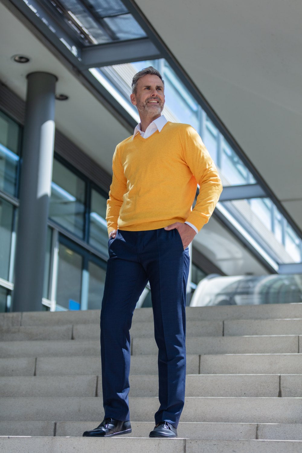 Ein Mann in gelbem Pullover und dunkler Hose steht selbstbewusst auf der Treppe vor einem modernen Gebäude, die Hände in den Taschen, und blickt in die Ferne. Seine schwarzen ZAQQ ARQ-Stiefel verleihen seinem Outfit einen Hauch von Eleganz.