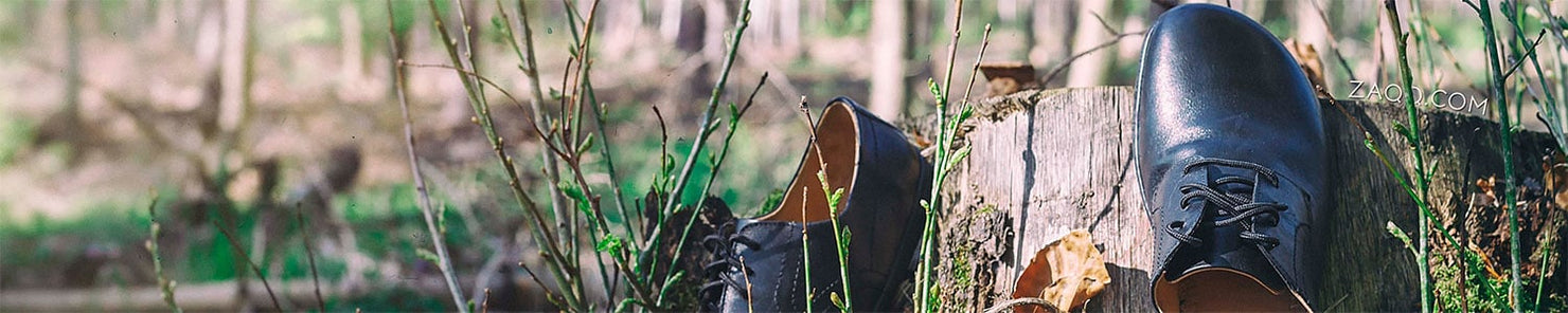 Nahaufnahme eines Paars schwarzer Lederschuhe, die auf einem Baumstumpf in einem Wald stehen. Die Schuhe haben an der Seite ein markantes Emblem. Im Hintergrund sind verschwommenes Grün und Bäume zu sehen, was eine ruhige Umgebung im Freien suggeriert.