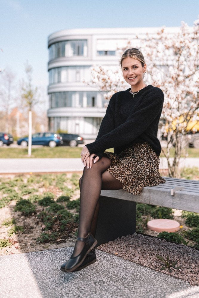 Eine Frau sitzt auf einer Bank im Freien. Sie trägt einen schwarzen Pullover, einen Rock mit Leopardenmuster und schwarze Strumpfhosen. Sie lächelt sanft, im Hintergrund sind ein modernes Gebäude und blühende Bäume zu sehen. Ihr Look wird durch elegante BANQET Nappa Dark Brown-Schuhe von ZAQQ vervollständigt.