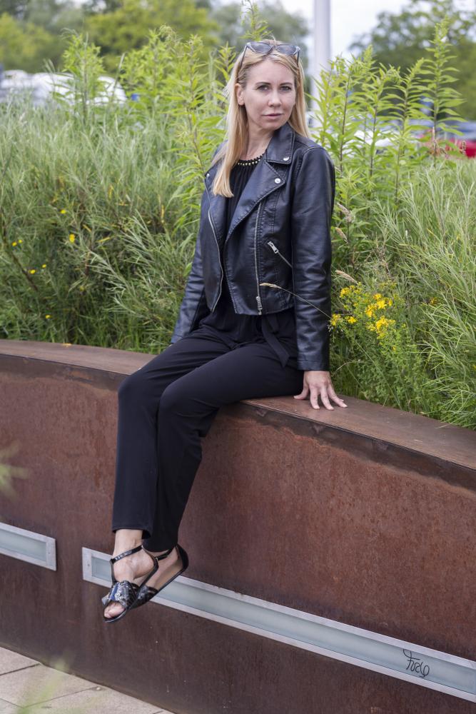 Eine Frau mit langen blonden Haaren sitzt auf einer großen rostfarbenen Metallbank. Sie trägt eine elegante schwarze Lederjacke, eine schwarze Hose und stylische BOUQET Black Sandalen von ZAQQ. Hinter ihr bilden hohe grüne Pflanzen eine üppige Kulisse.