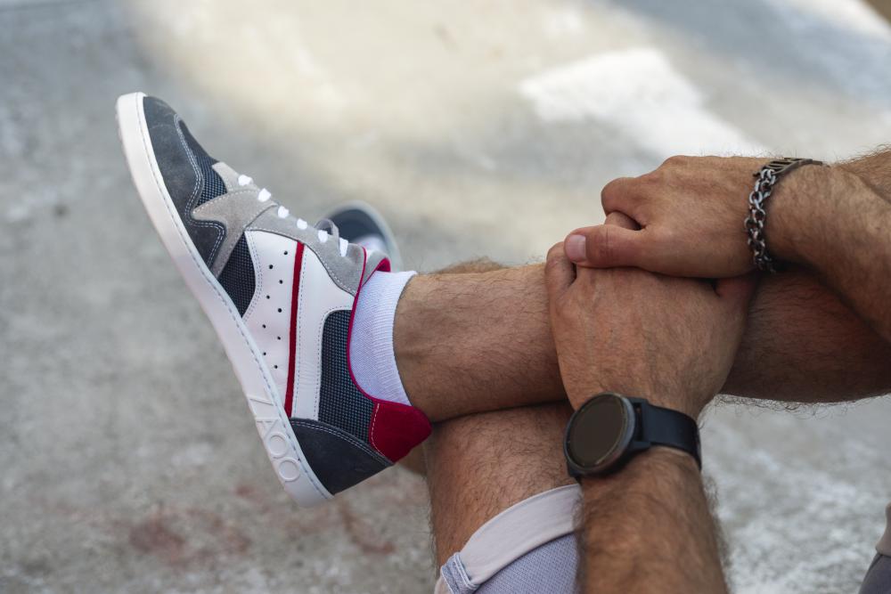 Eine Person sitzt mit gekreuzten Beinen da und hebt die BREAQ Darkblue-Sneaker von ZAQQ mit leuchtend roten, grauen und weißen Akzenten hervor. Ihre Hände sind über dem Knie gefaltet, geschmückt mit einem schwarzen Armband und einem silbernen Kettenarmband vor dem Hintergrund eines Betonbodens.