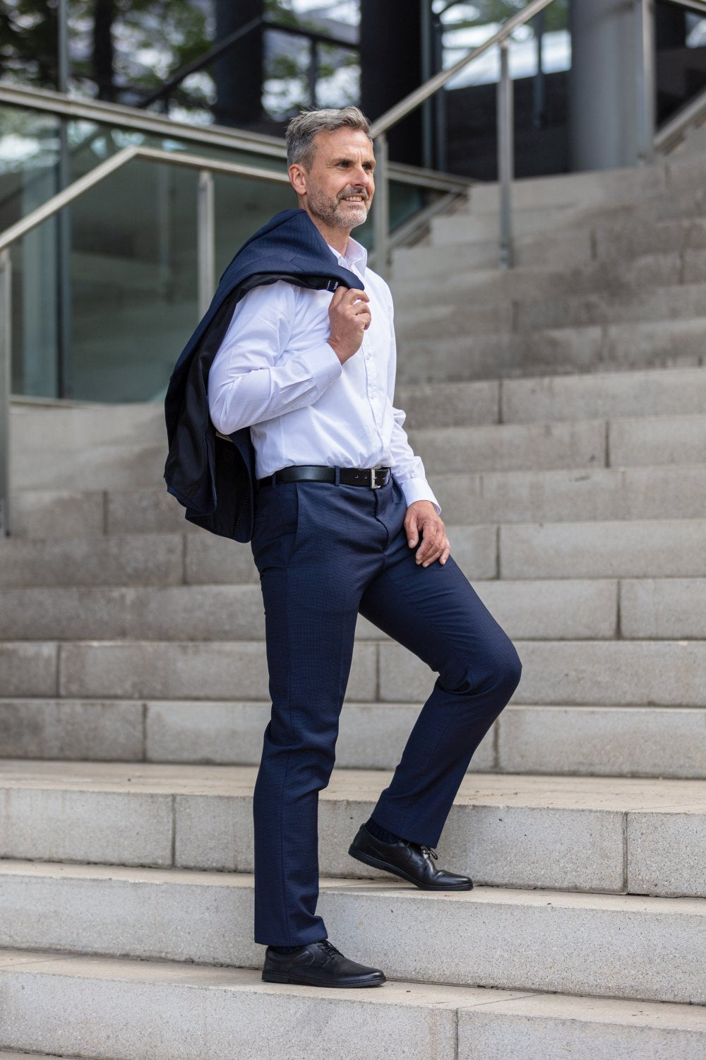 Ein Mann mit grauem Haar, gekleidet in ein weißes Hemd und eine dunkelblaue Hose, schreitet selbstbewusst die Außentreppe hinauf, mit einem Jackett über der Schulter. Seine zeitlose Eleganz wird durch die stilvollen BRIQ Black Business-Schuhe von ZAQQ vervollständigt, die perfekt zu seiner anspruchsvollen Kleidung passen.