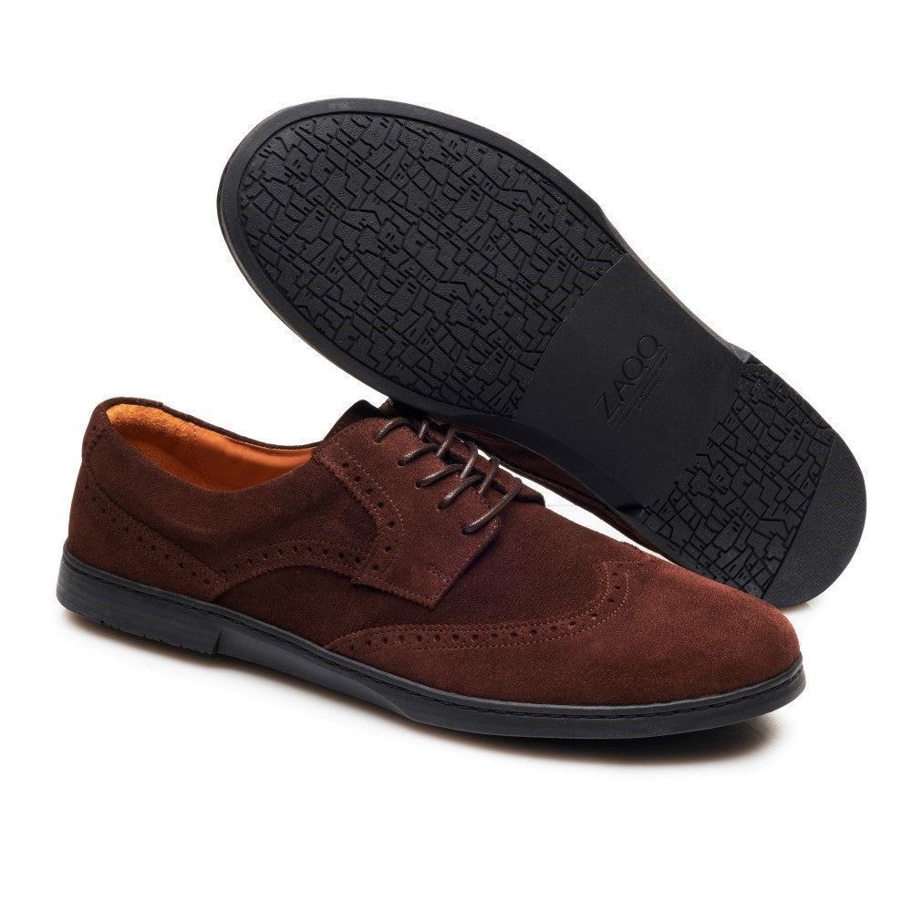 Der BRIQ Brogue Velours Brown von ZAQQ hat ein samtiges Finish mit Ziernähten und ein perforiertes Design. Diese Wildlederschuhe werden mit dunkelbraunen Schnürsenkeln geschnürt. Ein Schuh steht aufrecht, um sein Top-Design zu präsentieren, während der andere seine strukturierte schwarze Sohle hervorhebt.