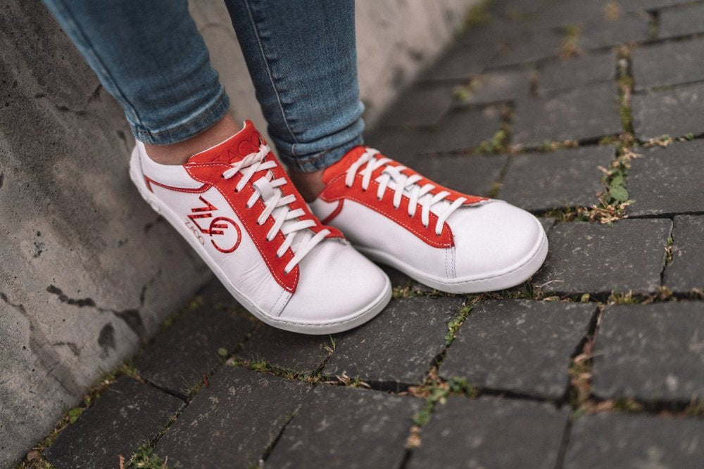 Eine Detailaufnahme einer Person, die die CEDRIQ Red-Sneaker von ZAQQ trägt, die das weiß-rote Design mit der Schrift „740“ auf der Seite zeigen, während sie auf einem strukturierten Kopfsteinpflaster steht. Der Sneaker ist mit einer flexiblen Kautschuksohle ausgestattet und wird modisch mit Blue Jeans kombiniert.