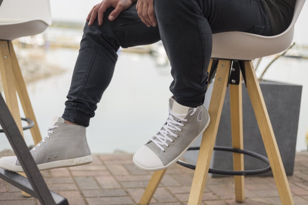 Zwei Personen sitzen auf hölzernen Barhockern, eine davon trägt ZAQQs graue High-Top-Sneakers CHUQQS aus Nappaleder und dazu schwarze Jeans. Die Außenszene entfaltet sich über einem Ziegelsteinpflaster und geht in einen verschwommenen Hintergrund aus Wasser und Booten über.