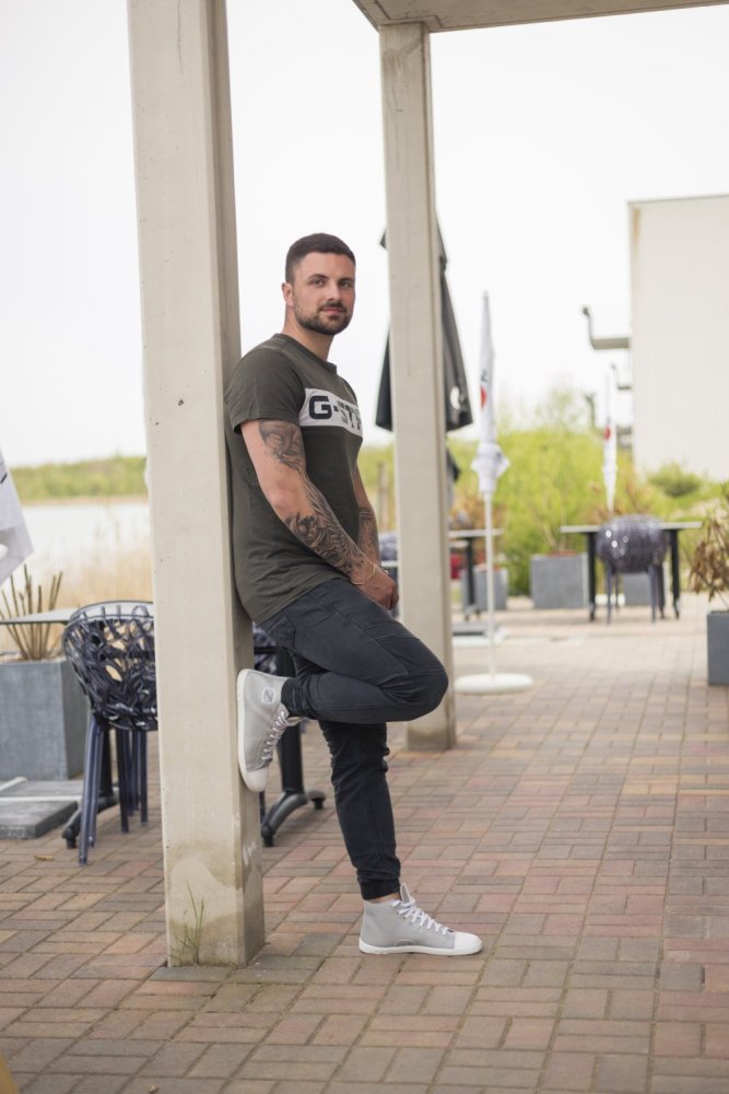 Ein Mann in grünem T-Shirt und schwarzer Hose lehnt an einem Betonpfeiler und präsentiert die grauen High-Top-Sneakers CHUQQS von ZAQQ. Hinter ihm ergänzen Stühle, Tische und üppiges Grün den hellen Himmel in dieser lebendigen Außenumgebung.