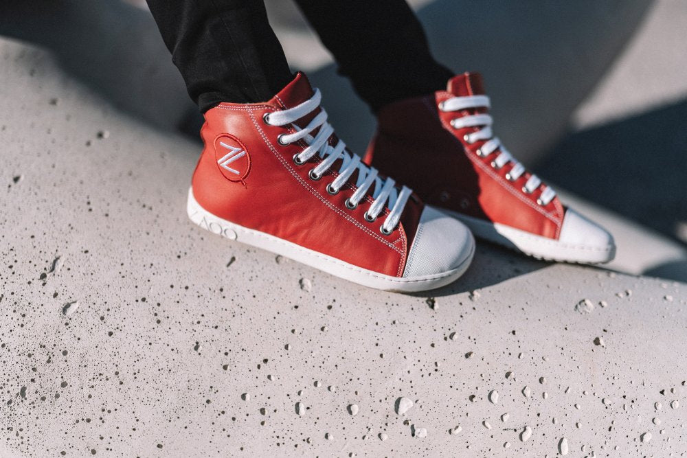 Ein Paar CHUQQS Red High-Top-Sneaker von ZAQQ, gefertigt aus geschmeidigem Nappaleder mit weißen Schnürsenkeln und Sohlen, ruhen auf einer strukturierten Betonoberfläche. Die Schuhe verfügen über ein kreisförmiges Logo an der Seite und vereinen Stil mit Komfort.