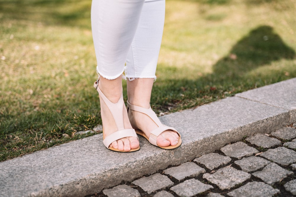 Eine Person in weißen Hosen und CLIQ Nude-Sandalen von ZAQQ steht am Rand eines Kopfsteinpflasterwegs mit Gras im Hintergrund.