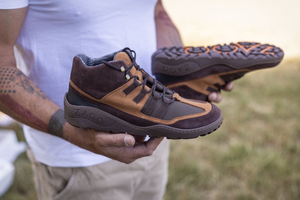 Eine Person in einem weißen T-Shirt hält ein Paar braune wasserdichte ESQAPE-Schuhe von ZAQQ mit robusten Sohlen, ideal für Fans des Barfußschuh-Designs. Die Mischung aus Wildleder und Mesh-Materialien ist ein Hingucker. Tattoos zieren den Arm der Person vor einem grasbewachsenen Hintergrund.