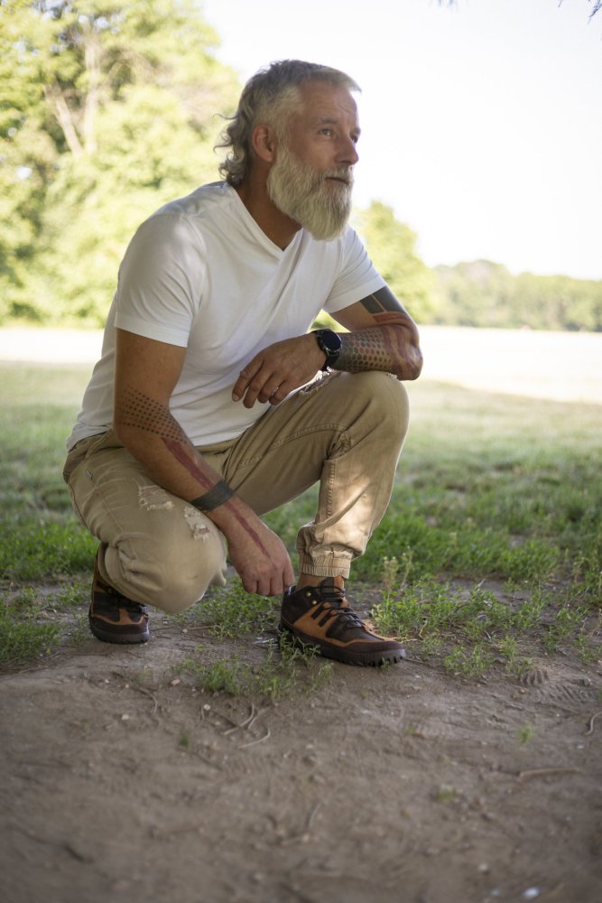 Ein Mann mit Bart, bekleidet mit einem weißen T-Shirt und Khakihosen, hockt auf einer Grasfläche im Freien. Er trägt die braunen wasserdichten ESQAPE-Schuhe von ZAQQ und zeigt Tattoos auf seinen Armen. Bäume und ein klarer Himmel bilden den Hintergrund und betonen seinen Abenteuergeist.