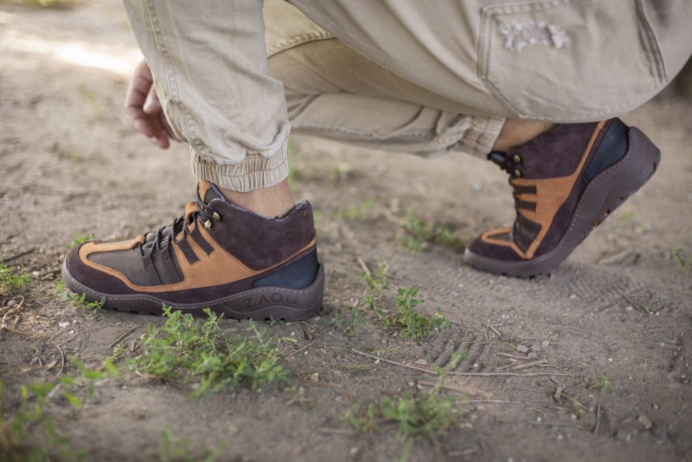 Eine Person hockt im Freien auf Erde und trägt braune wasserdichte Wanderschuhe von ZAQQ ESQAPE und hellbeige Hosen. Die Schuhe haben eine raue Textur, wodurch sie zum Wandern geeignet erscheinen. Aus dem Boden sprießen vereinzelt üppige grüne Pflanzen.