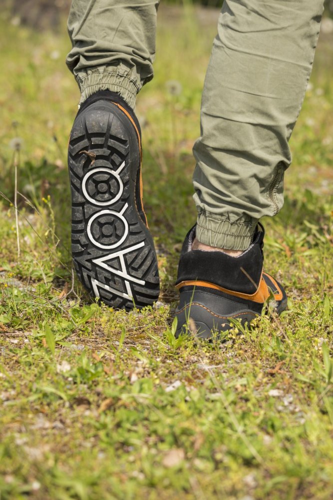 Eine Person läuft in braunen Wanderstiefeln und grünen Hosen mit einer wasserdichten Sohle in Schwarz-Orange von ESQAPE von ZAQQ auf Gras. Die Sohle des linken Stiefels ist sichtbar und weist markante schwarz-weiße Muster und kreisförmige Designs auf. Der Grasboden ist mit kleinen Pflanzen und Blumen gesprenkelt.