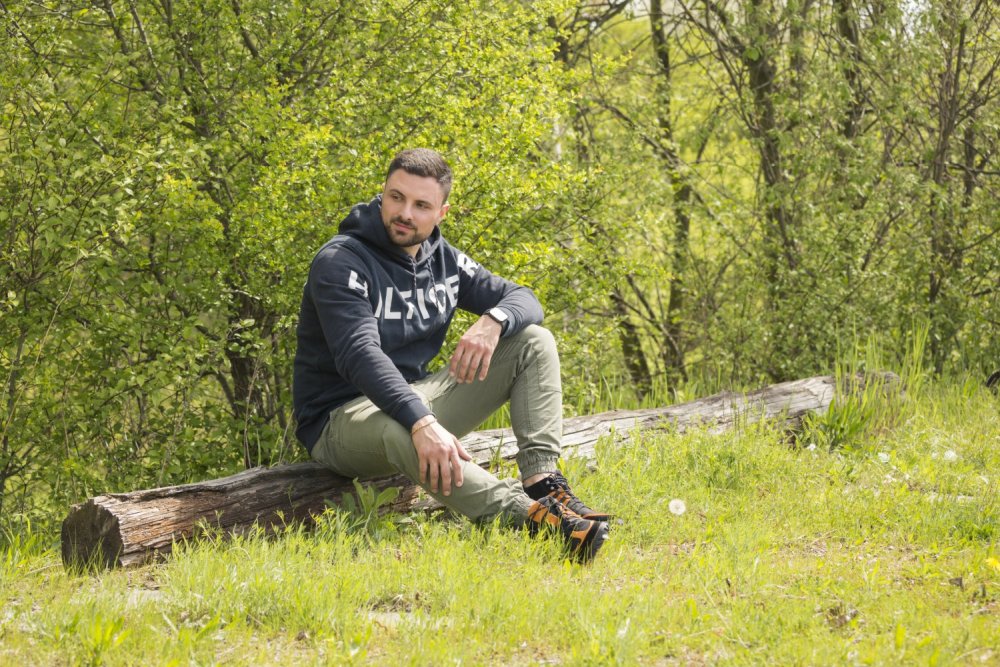 Ein Mann sitzt auf einem umgestürzten Baumstamm in einer üppigen, grünen Waldlandschaft. Er trägt einen wasserdichten ESQAPE Black Orange Hoodie von ZAQQ und hellgrüne Hosen und blickt in die Ferne. Der Hintergrund ist voller belaubter Bäume und Gras und bietet eine ruhige Flucht vor der alltäglichen Hektik.