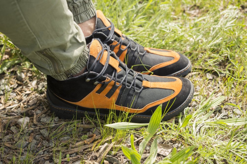 Eine Nahaufnahme einer Person, die die wasserdichten ESQAPE Black Orange-Wanderstiefel von ZAQQ trägt und auf einer Grasfläche mit kleinen Pflanzen und Kies steht. Die Stiefel haben detaillierte Nähte und dicke Sohlen und ergänzen die grüne Hose perfekt.