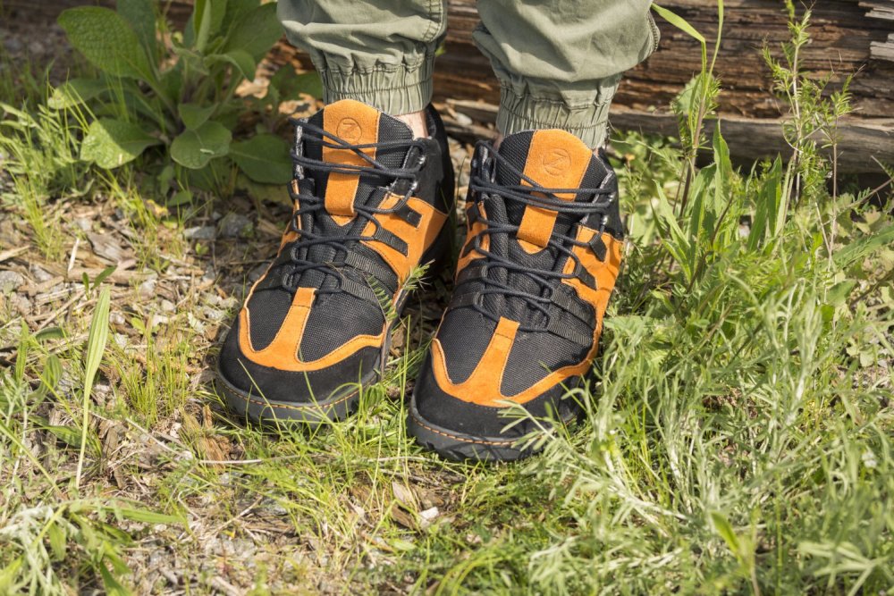 Eine Person, die die stylischen ESQAPE Black Orange Waterproof High-Top-Sneaker mit ZAQQ TERRA-Sohle trägt, steht auf Gras und Kies, umgeben von üppigen grünen Pflanzen und einem Holzstamm. Ihre hellgrüne Hose ist an den Knöcheln stylisch gerafft.