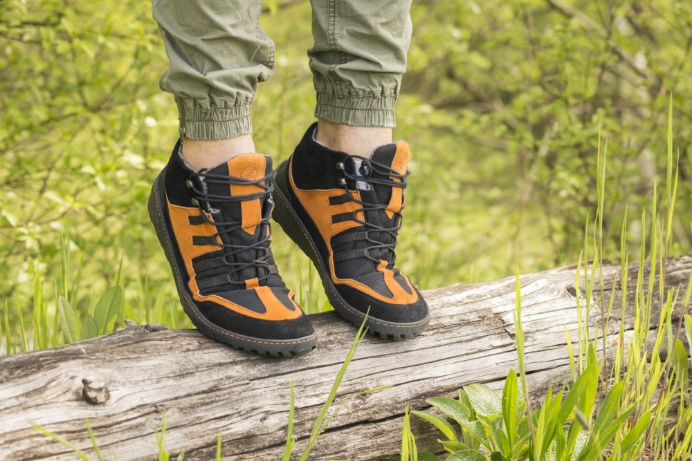 Eine Person in den wasserdichten ESQAPE-Stiefeln in Schwarz und Orange von ZAQQ steht selbstbewusst auf einem umgestürzten Baumstamm inmitten von üppigem Grün. Sie trägt hellgrüne Hosen, die die natürliche Atmosphäre im Freien unterstreichen.