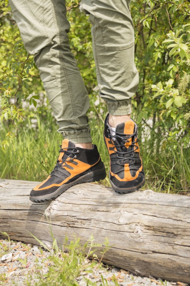 Eine Person in grünen Cargohosen und wasserdichten schwarz-orangefarbenen Schuhen von ZAQQ ESQAPE steht auf einem Baumstamm in einer grasbewachsenen Außenumgebung, umgeben von üppigem grünem Laub.