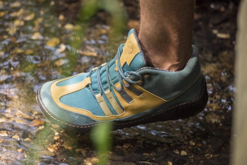 Wer die wasserdichten Schuhe ESQAPE Blue Orange von ZAQQ trägt, kann selbstbewusst in einen seichten Bach steigen, denn ihr wasserdichtes Design garantiert trockenen Komfort. Unter dem klaren Wasser sind Steine zu sehen, inmitten einer üppigen und grünen Naturlandschaft.