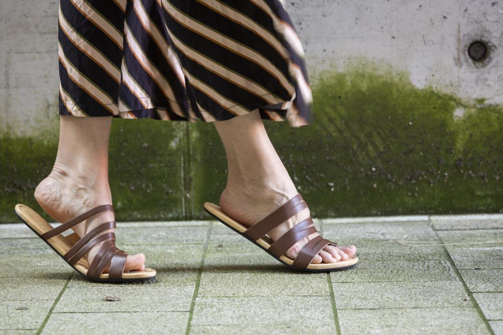 Eine Person schlendert einen grün gefliesten Weg entlang und trägt die FLAQE Dark Brown Barfußsandalen von ZAQQ und einen langen, gestreiften Rock. Im Hintergrund trägt eine Betonwand mit Moos zur Atmosphäre bei und unterstreicht sowohl Stil als auch außergewöhnlichen Komfort.