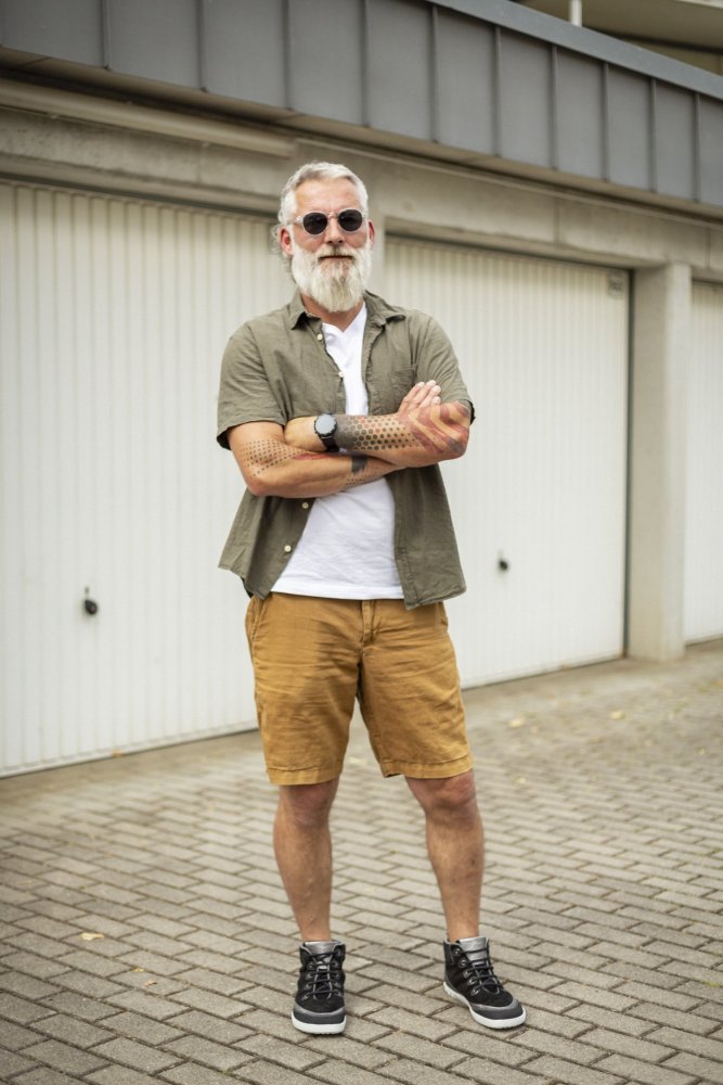 Ein bärtiger Mann mit Sonnenbrille steht selbstbewusst draußen, gekleidet in ein olivgrünes Hemd über einem weißen T-Shirt, dazu gelbe Shorts und die schicken schwarzen FRANQY-Sneaker von ZAQQ. Er hat die Arme verschränkt und posiert vor einer Reihe geschlossener Garagentore.