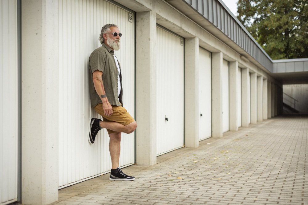 Ein Mann mit Sonnenbrille und Bart lehnt an einer Reihe von Garagentoren, mühelos stilvoll in seinem grünen Hemd, weißem T-Shirt und braunen Shorts. Ein Fuß ruht auf der Wand hinter ihm und zeigt die eleganten schwarzen ZAQQ FRANQY-Sneaker. Der gepflasterte Boden kontrastiert mit den üppigen Bäumen im Hintergrund.