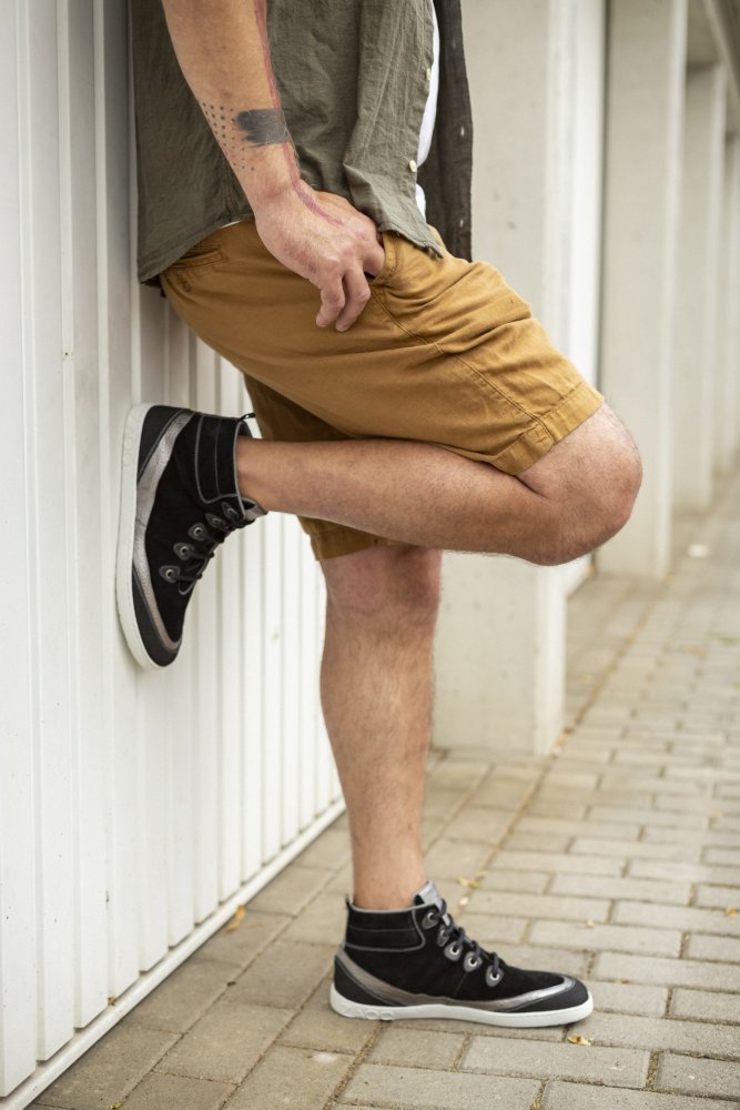 Eine Person lehnt mit angewinkeltem Bein an einer weißen Wand. Sie trägt ein grünes Shirt, braune Shorts und Midcut-Sneaker aus der ZAQQ-Kollektion namens FRANQY Black. Im Mittelpunkt stehen das lässige Outfit und die auffälligen Armtattoos. Das Setting ist ein mit Ziegeln gepflasterter Boden.