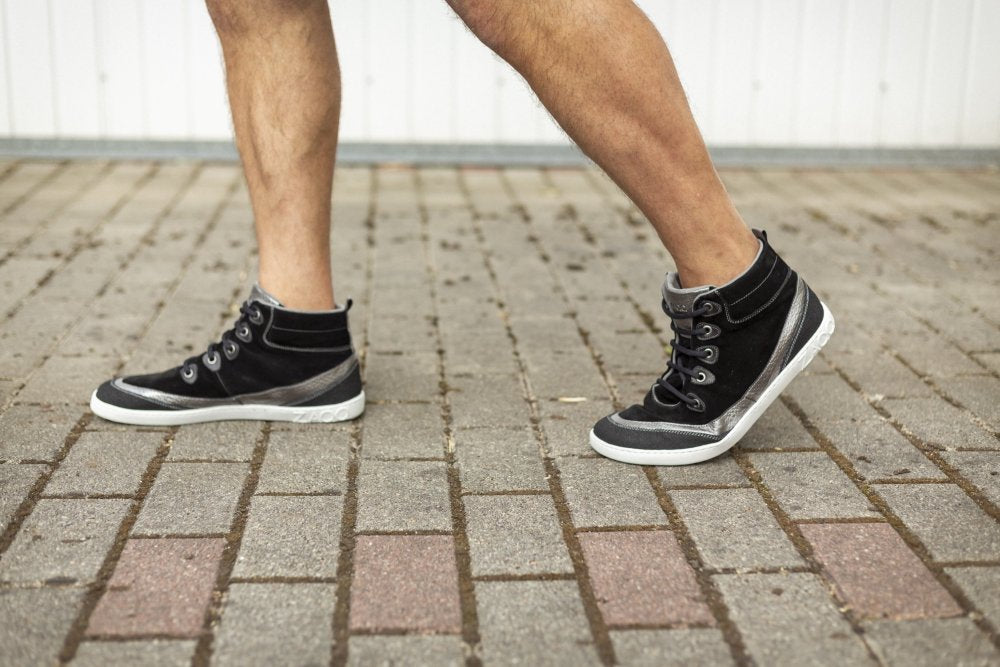 Eine Person schlendert auf einem Ziegelweg und trägt stylische schwarze Midcut-Sneaker von ZAQQ FRANQY. Vor einer strahlend weißen Wand im Hintergrund sind nur die Unterschenkel und Füße zu sehen.