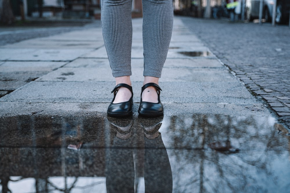 Eine Person in karierten Hosen und ZAQQs BANQET Nappa Black Mary Jane Schuhen steht vor einer Pfütze auf einem gepflasterten Weg. Im Wasser ist die Spiegelung ihrer Beine und Schuhe zu sehen. Der Weg führt in einen verschwommenen urbanen Hintergrund.