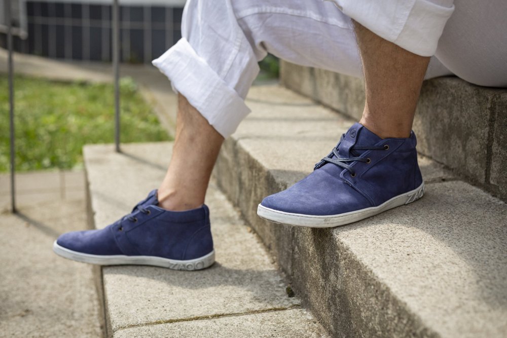 Eine Person in hochgekrempelten weißen Hosen sitzt auf Betonstufen und präsentiert ZAQQs LANQASTER Navy-Schuhe mit weißen Sohlen. In der Außenszene ist im Hintergrund Gras zu sehen.