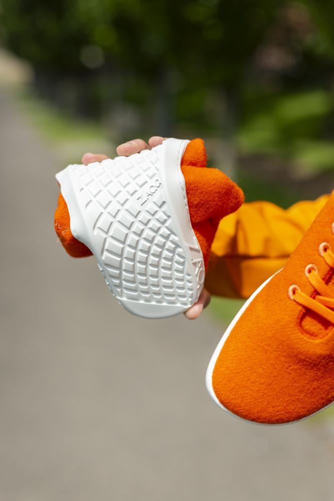 Eine Person hält den orangefarbenen Schuh ZAQQ LIQE mit flexibler weißer Sohle und biegt ihn, um sein Barfußgefühl zu demonstrieren. Der Schuh hat orangefarbene Schnürsenkel und wird mit einem passenden orangefarbenen Ärmel gehalten, der vor einem verschwommenen Außenhintergrund steht.