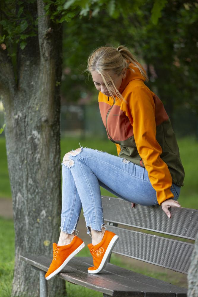 Eine Person sitzt auf einer Parkbank, gekleidet in eine orange-grüne Jacke, zerrissene Jeans und LIQE Orange Sneakers von ZAQQ. Sie strahlt ein Gefühl von Barfuß aus, während sie nach unten blickt, während im Hintergrund ein Baum und Grün sanft für die natürliche Temperaturregulierung sorgen.