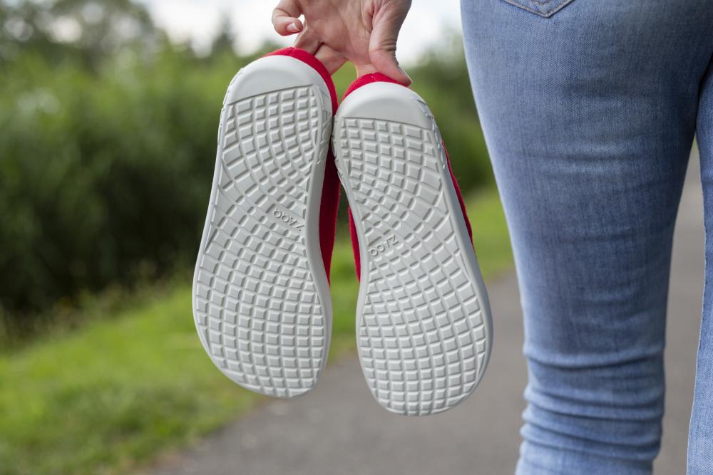 Eine Person in Blue Jeans hält ein Paar LIQE Race-Sneaker an den Fersen und zeigt ihre weißen Sohlen mit einem Gittermuster. Sie steht auf einem Weg, der von grünem Gras und verschwommenen Bäumen im Hintergrund umgeben ist.