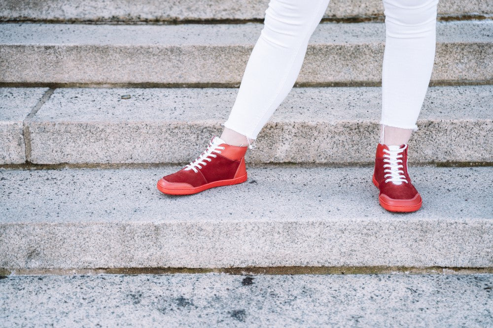 Eine Person in weißer Hose steht selbstbewusst auf Betonstufen und trägt die stylischen LUQQ Red High-Top Sneaker von ZAQQ mit flexibler Kautschuksohle. Der Hintergrund besteht aus Steinstufen, der Fokus liegt jedoch klar auf dem schicken Schuhwerk.