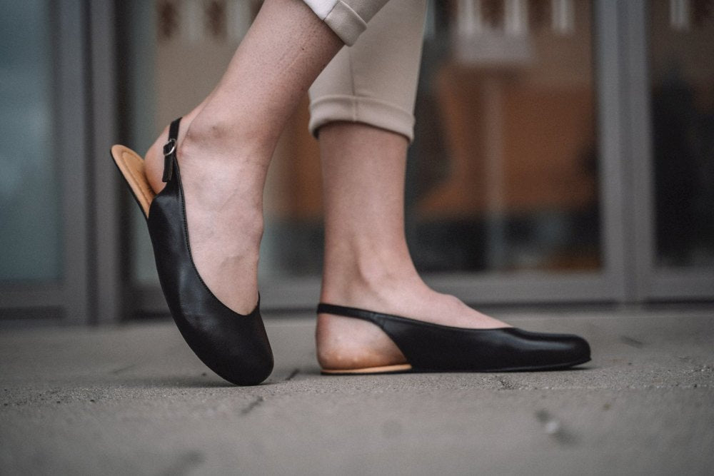 Eine Person in beigefarbenen Hosen und schwarzen PINQ-Schuhen von ZAQQ, die den Komfort einer Ballerina verkörpert, steht auf einer Betonfläche. Der Hintergrund ist verschwommen und zeigt Glastüren und eine Gebäudeaußenseite.