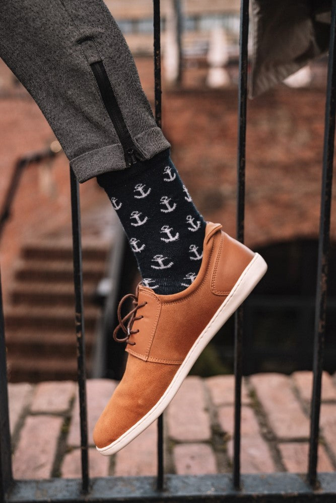 Nahaufnahme einer Person, die die PIQUANT Sierra Sneaker von ZAQQ in Braun trägt, gepaart mit schwarzen Socken mit weißem Ankermuster, und auf eine Ziegeloberfläche tritt. Die Sneaker sind aus Premium Veloursleder gefertigt und bieten so mehr Komfort. Die Person trägt eine dunkelgraue Hose mit seitlichem Reißverschluss und im Hintergrund ist ein schmiedeeisernes Geländer zu sehen.